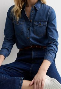 Person som sitter i skräddarställning iförd en blå denimskjorta och jeans med ett brunt bälte och vita sneakers, mot en enfärgad bakgrund.