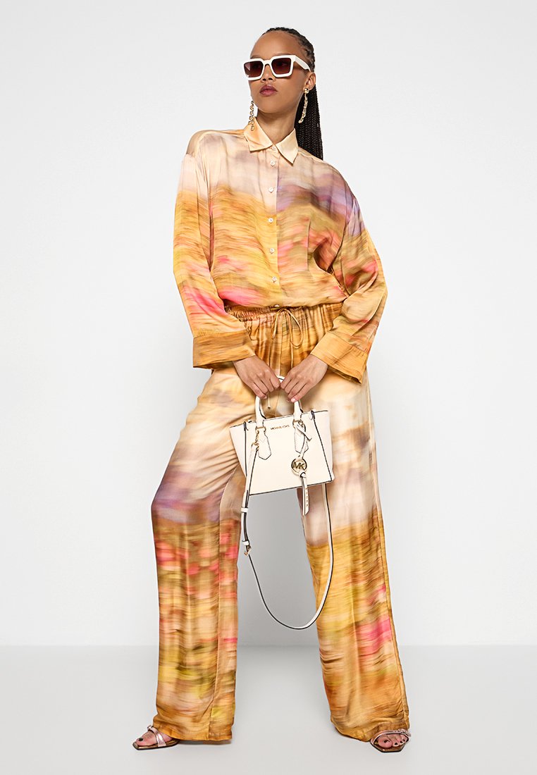 Frau mit geflochtenem Haar trägt einen übergroßen gelb-pink gemusterten Anzug, weiße Sonnenbrille, goldene Ohrringe und hält eine weiße Handtasche.