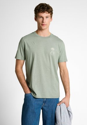 Młody mężczyzna w jasnozielonej koszulce z grafiką palmy, niebieskich dżinsach, trzyma w prawej ręce koszulę w paski, stoi na białym tle.