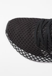 Czarny sportowy but z siateczkową cholewką o fakturowanej powierzchni, okrągłym noskiem i wytrzymałą podeszwą z odblaskowym akcentem po bokach. Widoczne sznurowadła do regulacji.