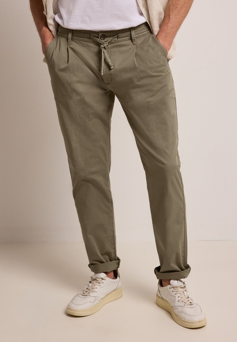 Homme portant un pantalon kaki retroussé avec taille à cordon, baskets blanches, mains dans les poches, debout sur un sol et un fond neutres.