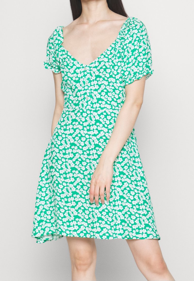Femme portant une robe verte avec un imprimé floral blanc, des manches courtes à volants, un décolleté en V et des boutons sur le devant.