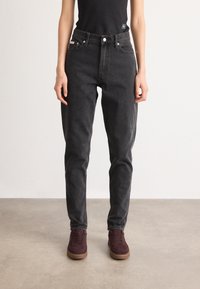 Person med høyde i sort jeans med høy midje, svart ermeløs topp og burgunder joggesko som står på et grått gulv mot en enkel hvit vegg.