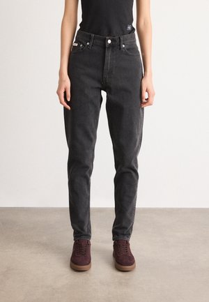 Persoon in hoge zwarte jeans, zwart mouwloos topje en bordeauxrode sneakers staat op een grijze vloer tegen een witte muur.