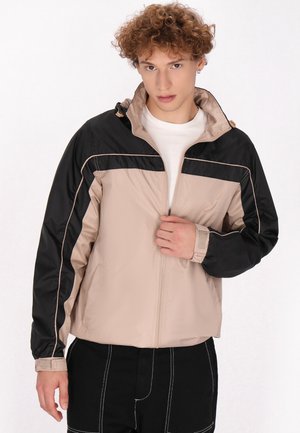 Beige en zwarte windbreaker jas met een hoge kraag, ritsluiting, elastische manchetten en contrasterende stikdetails langs de naden.