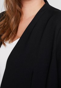 Mujer con cabello largo castaño que lleva un blazer negro sobre una blusa blanca, mostrada desde el cuello hasta la parte superior del pecho sobre un fondo sencillo.