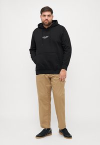 Zwarte capuchontrui met een voorzak en trekkoorden, gecombineerd met beige broeken en zwarte sneakers. Eenvoudig, minimalistisch ontwerp.