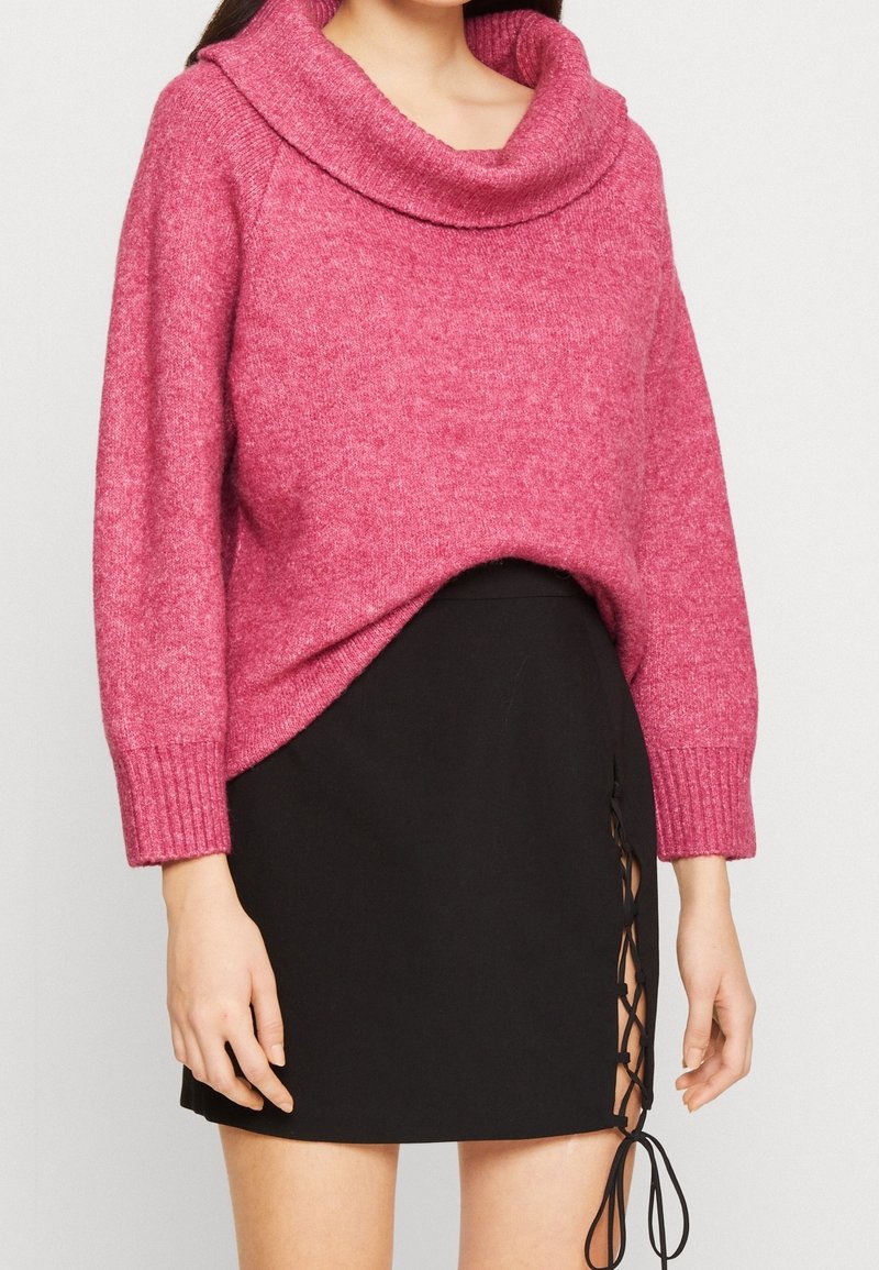 Femme portant un pull en maille rose avec un col large et une mini-jupe noire avec un détail de laçage sur le côté.