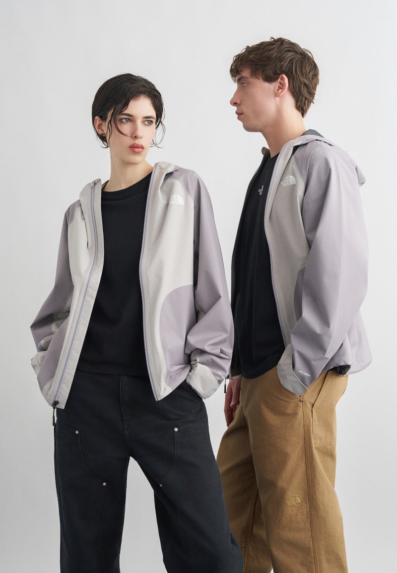 Δύο μοντέλα φορούν ανοιχτό γκρι μπουφάν North Face πάνω από μαύρα τοπ, το ένα κοιτάει μπροστά με τα χέρια στις τσέπες, το άλλο προφίλ.