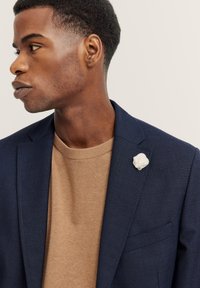 Blazer blu navy con finitura testurizzata, abbinato a un maglione beige. Un fermaglio a forma di fiore in tessuto bianco accentua il risvolto del blazer.