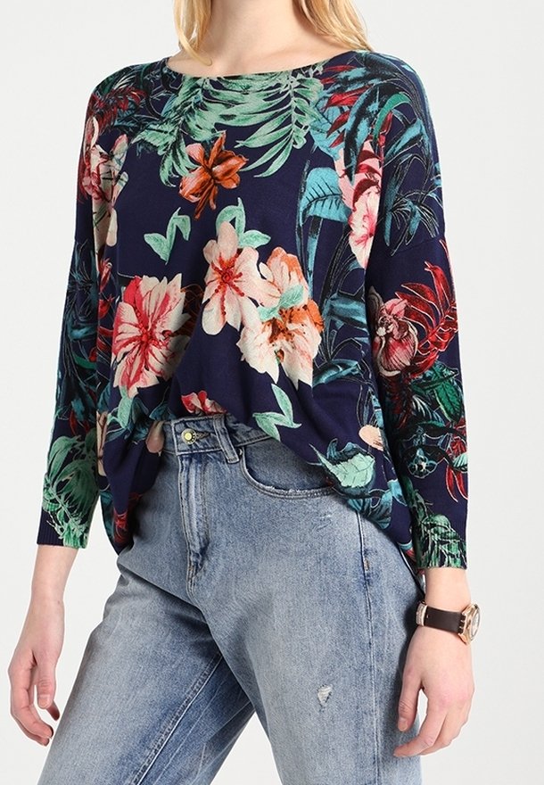 Femme portant un haut à manches longues floral bleu foncé associé à un jean taille haute bleu clair et une montre-bracelet.