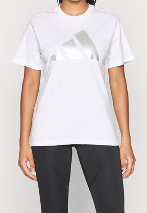 Kobieta ubrana w białą koszulkę z dużym srebrnym logo Adidas oraz czarne legginsy, stojąca na tle jednolitego jasnego tła.