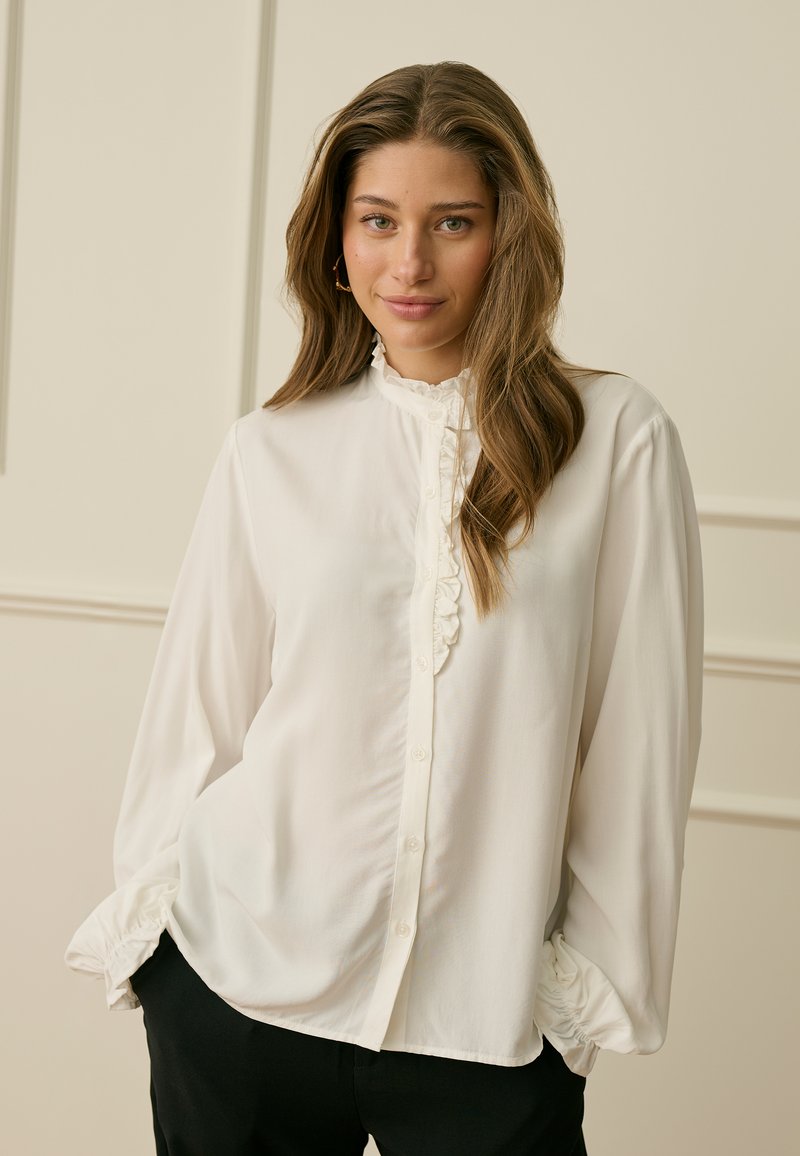 Mlada ženska z dolgimi rjavimi lasmi, oblečena v belo bluzo z gumbi in volančastim ovratnikom ter črne hlače, stoji pred svetlo steno.