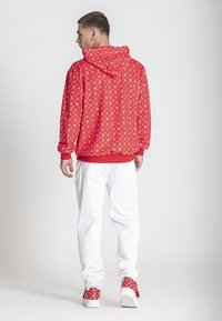 Sudadera roja con patrón de logo blanco, capucha y puños acanalados, combinada con pantalones blancos y zapatillas con detalles rojos. La vista trasera muestra los detalles del diseño.