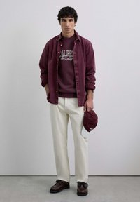 Camisa de botones color burdeos sobre un sudadera a juego, pantalones crema, zapatos marrón oscuro y una gorra burdeos con acentos blancos en la mano.