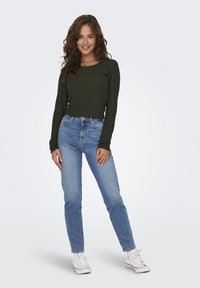 Mörkgrön ribbad långärmad topp som parats med blå högmidjade jeans och vita sneakers. Modellen står centrerad med en neutral bakgrund.