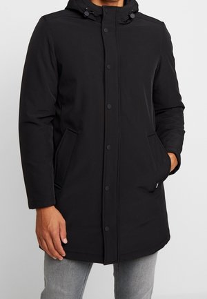 Hombre con un abrigo de invierno negro con capucha, botones a presión y bolsillos laterales, combinado con pantalones gris claro, de pie frente a un fondo blanco.