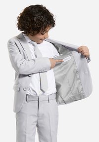 Giacca di suit grigio chiaro con rever a metà, due bottoni e interno in satin. Il bambino punta a un'etichetta con "OPPOSUITS" all'interno della giacca.