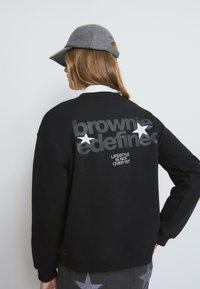 Sweatshirt noir avec un texte gris métallique et des étoiles blanches au dos, doté de poignets et d'un ourlet côtelés. Associé à une casquette grise.
