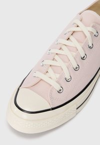 Zapatillas de lona rosas con punta de goma crema y ribete negro en la suela. Presentan cordones blancos y ojales metálicos. Diseño simple y bajo.