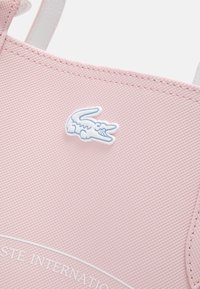 Tissu rose texturé avec un petit logo crocodile blanc et bleu clair centré, bords cousus visibles en haut et à droite.