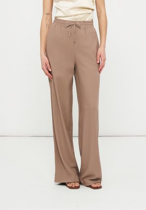 Kvinde iført lysebrune vidde-bukser med elastik i taljen og snøre, kombineret med åbne sandaler, foran en ensfarvet baggrund.