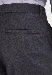 Pantaloni grigio scuro realizzati in tessuto strutturato con una sola tasca posteriore. La vita è piatta con una finitura pulita e dettagli di cucitura discreti.