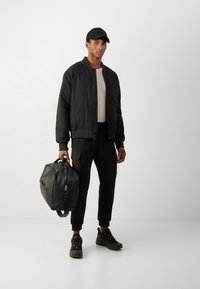 Svart bomberjakke med glatt finish, kremfarget t-skjorte, svarte joggebukser, svarte sneakers og en stor svart duffelbag.