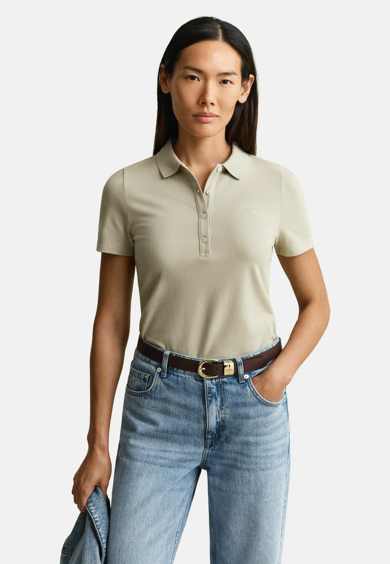 Femme portant un polo beige clair à manches courtes, rentré dans un jean bleu avec une ceinture marron, tenant une veste en denim dans une main.