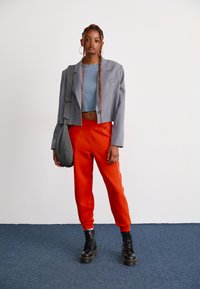 Jonge vrouw met lange ingevlochten haren, gekleed in een grijze cropped blazer, een blauwe top, feloranje broek, zwarte laarzen en een geruite tas.