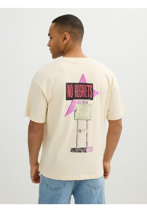 Hombre mirando hacia atrás, lleva una camiseta beige con el texto "NO REGRETS" sobre una estrella rosa y un gráfico de un cartel vintage de motel en la espalda.
