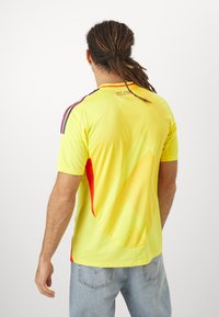 Yellow soccer jersey with red accents, short sleeves, and textured pattern. Features gradient design and contrasting shoulder stripes.