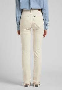 Jeans beige claro de mezclilla, con forma ajustada, bolsillos traseros y detalles de costura sutiles, usados con una camisa de color azul claro.