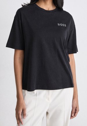 Femme portant un t-shirt noir ample avec « BOSS » imprimé sur la poitrine, associé à un pantalon blanc, debout sur un fond uni.