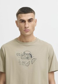 Beige cotton t-shirt featuring a graphic print of a tin can, fish, and text. Short sleeves and a crew neck design.