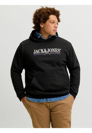 Junger Mann mit lockigem Haar, der einen schwarzen Jack & Jones Hoodie, blaues Hemd und beigefarbene Hose trägt, schaut nach rechts und hat die Hände in den Taschen.