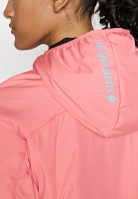 Vrouw met krullend haar die een koralen roze jas met capuchon en een reflecterend merkje op de achterkant draagt.