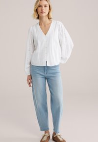 Blouse en coton blanc avec un col en V, une patte de boutonnage à l'avant et des manches longues bouffantes texturées ornées de broderies ajourées. Associée à un jean bleu clair.