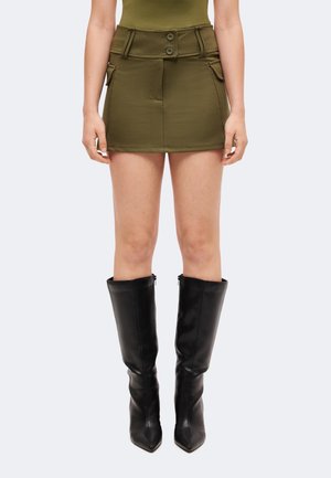 Mini-jupe vert olive avec une large ceinture et des boutons, portée avec des bottes en cuir noires montantes jusqu'aux genoux, à bout pointu.