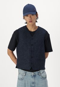 Marineblå ullblandingsvest med V-hals, seks svarte knapper og to frontlommer, brukt over en svart T-skjorte og lyseblå jeans.