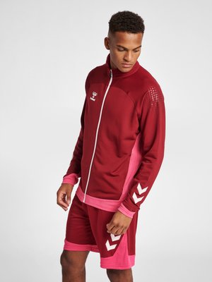 Rote Sportjacke mit pinken Akzenten und Reißverschluss, gemusterte Schultern, kombiniert mit passenden roten Shorts mit weißen Chevron-Details.