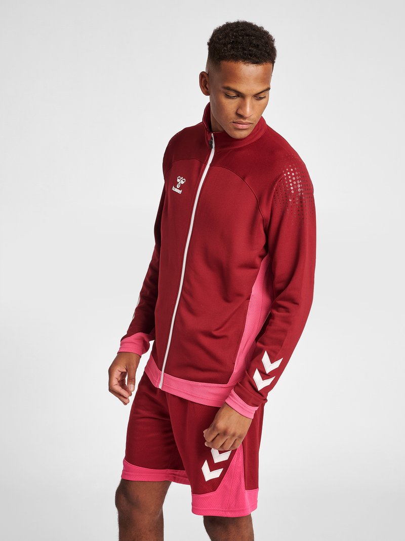 Rød atletisk jakke med pink detaljer og lynlås, mønstrede skuldre, parret med matchende røde shorts med hvide chevron-detaljer.