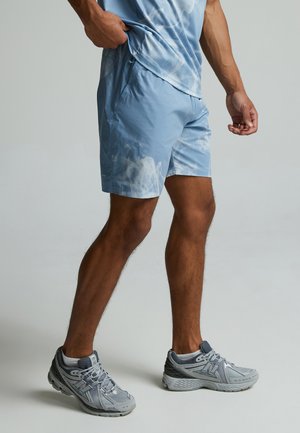 Homme portant un short tie-dye bleu clair, une chemise assortie et des baskets New Balance grises, debout avec une main dans la poche.