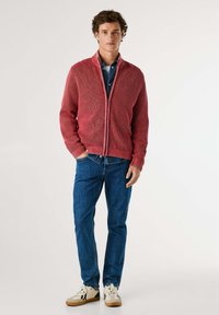 Rød strikket zip-up sweater med ribbet tekstur, båret over en blå denimskjorte, parret med blå jeans og hvide sneakers.