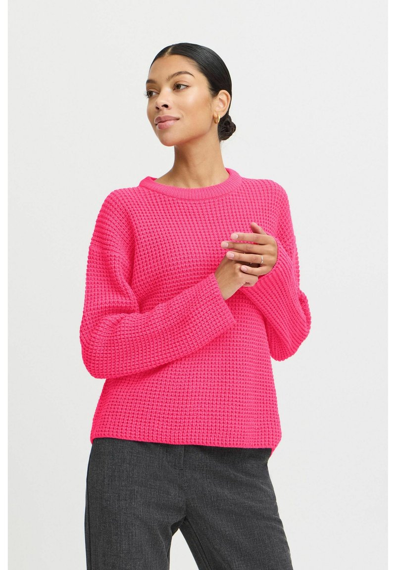 Pull en tricot rose vif avec un motif texturé, col rond et coupe décontractée, associé à un pantalon gris foncé.