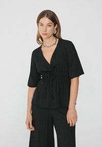 Schwarze, strukturierte Bluse mit tiefem V-Ausschnitt, kurzen Ärmeln und einer Kordelzugbindung in der Taille. Kombiniert mit passenden weit geschnittenen Hosen.