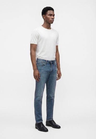 Homme debout portant un t-shirt blanc uni à manches courtes, un jean bleu et des chaussures noires habillées sur un fond blanc uni.