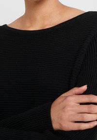 Pull en tricot noir avec une texture côtelée, un large col bateau et des manches longues. Vue rapprochée du tissu et des bras croisés.