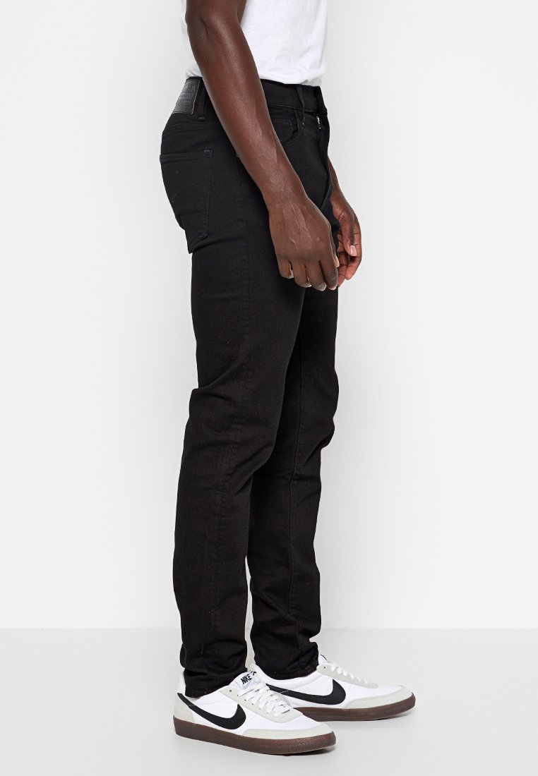 Homme portant un slim noir, un T-shirt blanc et des baskets Nike blanches avec un swoosh noir, se tenant de côté devant un fond blanc.