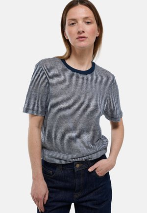 Femme aux cheveux courts bruns portant un t-shirt à manches courtes rayé bleu marine et blanc et un jean bleu foncé avec une main dans la poche.